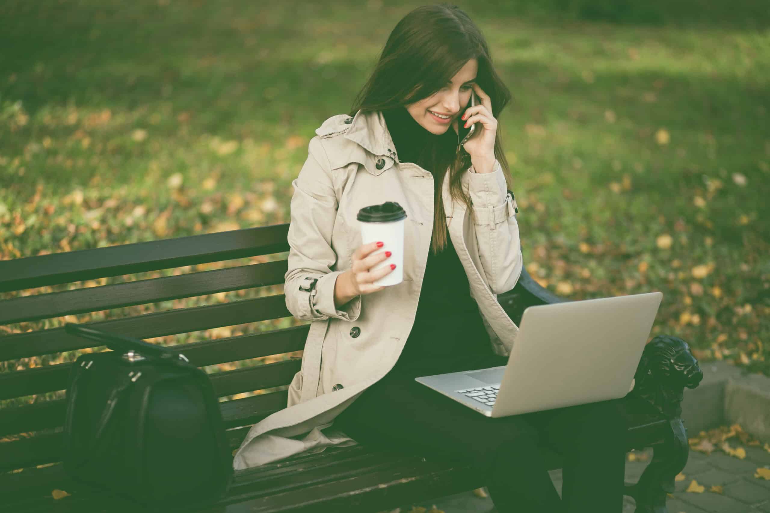 Una chica sentada en un banco de parque, con un portátil sobre sus piernas. Sostiene un café en una mano mientras atiende una llamada con la otra.