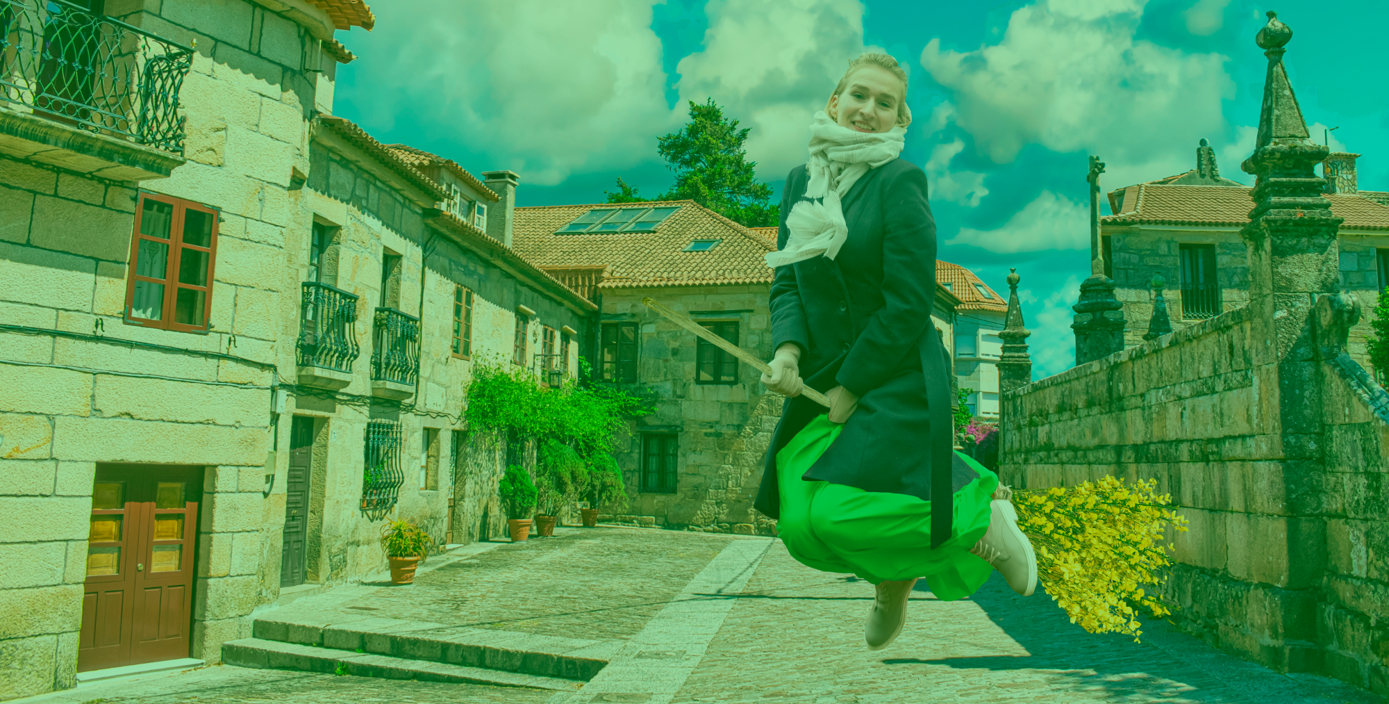 Chica subida en una escoba simulando un vuelo, entre un paisaje de casas de piedra.